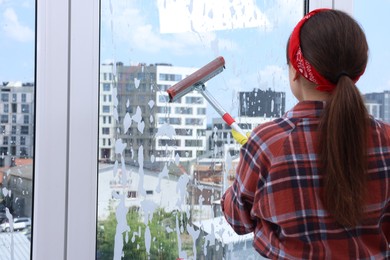 Photo of Woman with squeegee tool and foam cleaning window indoors, back view