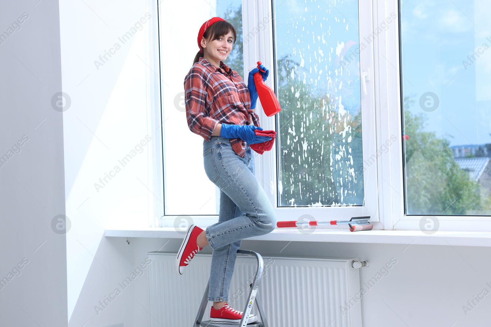 Photo of Beautiful young woman with spray bottle of detergent and napkin cleaning window indoors