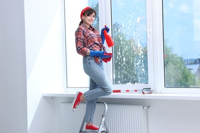 Beautiful young woman with spray bottle of detergent and napkin cleaning window indoors