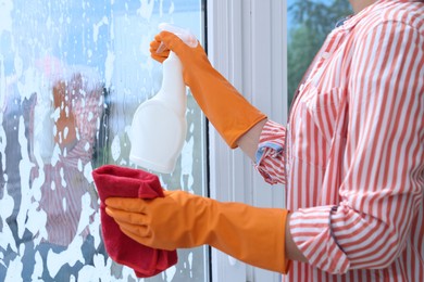 Woman with spray bottle of detergent and napkin cleaning window indoors, closeup