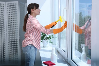 Beautiful young housewife with squeegee cleaning window indoors