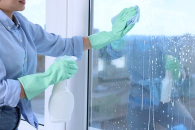 Woman with spray bottle of detergent and napkin cleaning window indoors, closeup