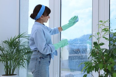 Young woman with spray bottle of detergent and napkin cleaning window indoors