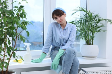 Photo of Cleaning window. Tired young woman with spray bottle of detergent and napkin on windowsill indoors