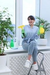 Beautiful young woman with squeegee, spray bottles of detergent and napkin on windowsill indoors