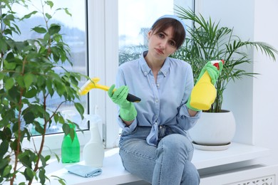 Tired young woman with squeegee, spray bottles of detergent and napkin on windowsill indoors