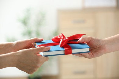 Woman gifting her friend book tied with red ribbon indoors, closeup