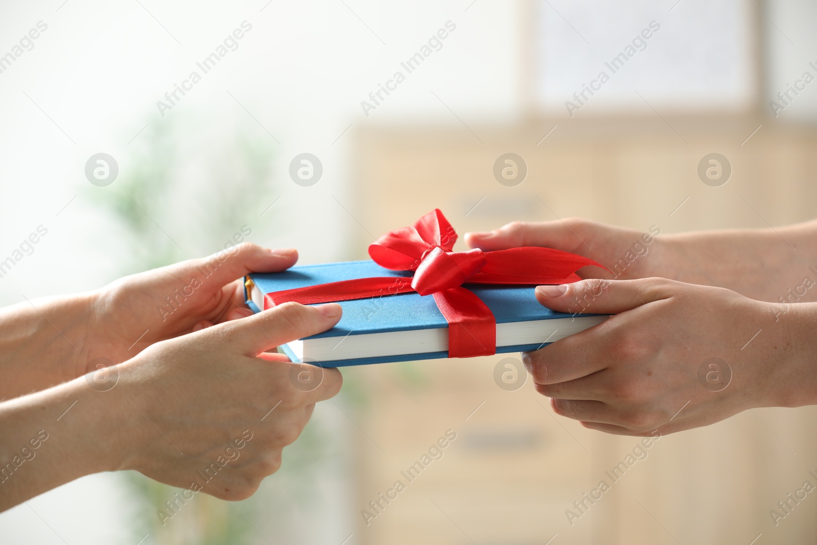 Photo of Woman gifting her friend book tied with red ribbon indoors, closeup