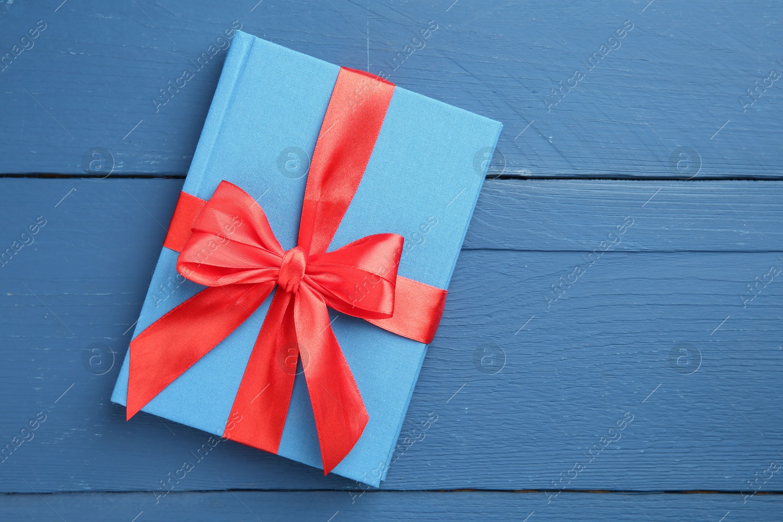 Photo of Book tied with red ribbon as gift on blue wooden table, top view. Space for text