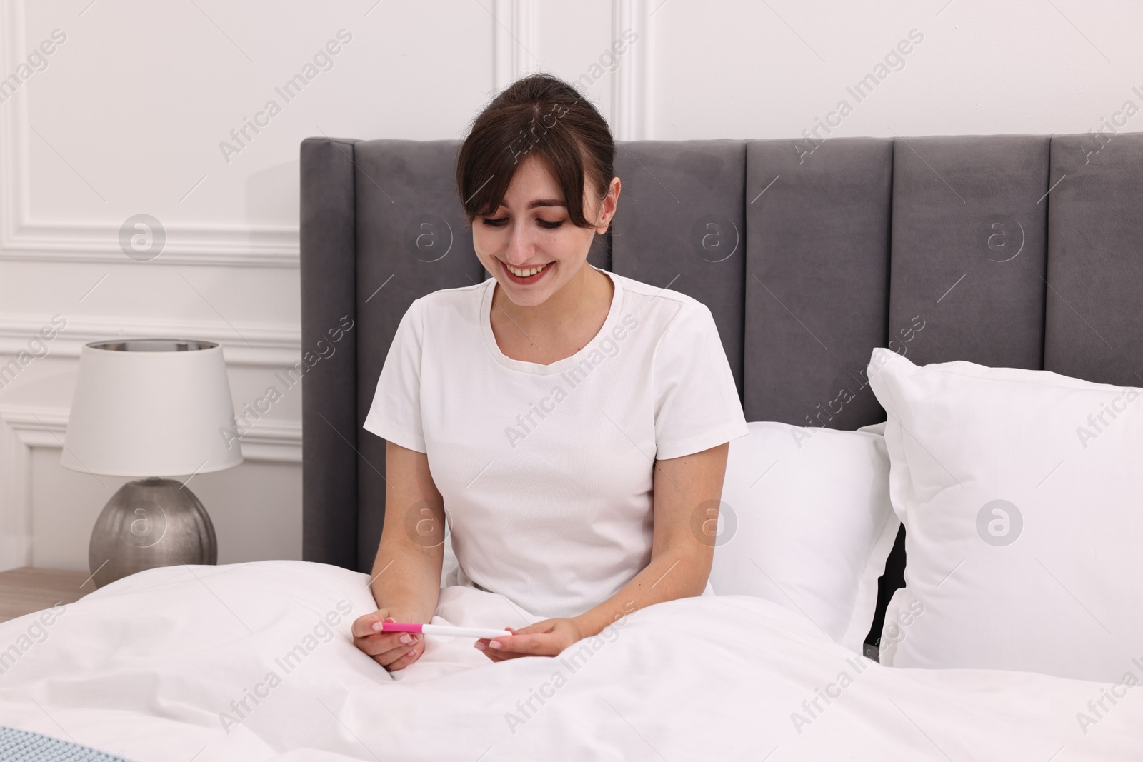 Photo of Happy young woman with pregnancy test on bed at home