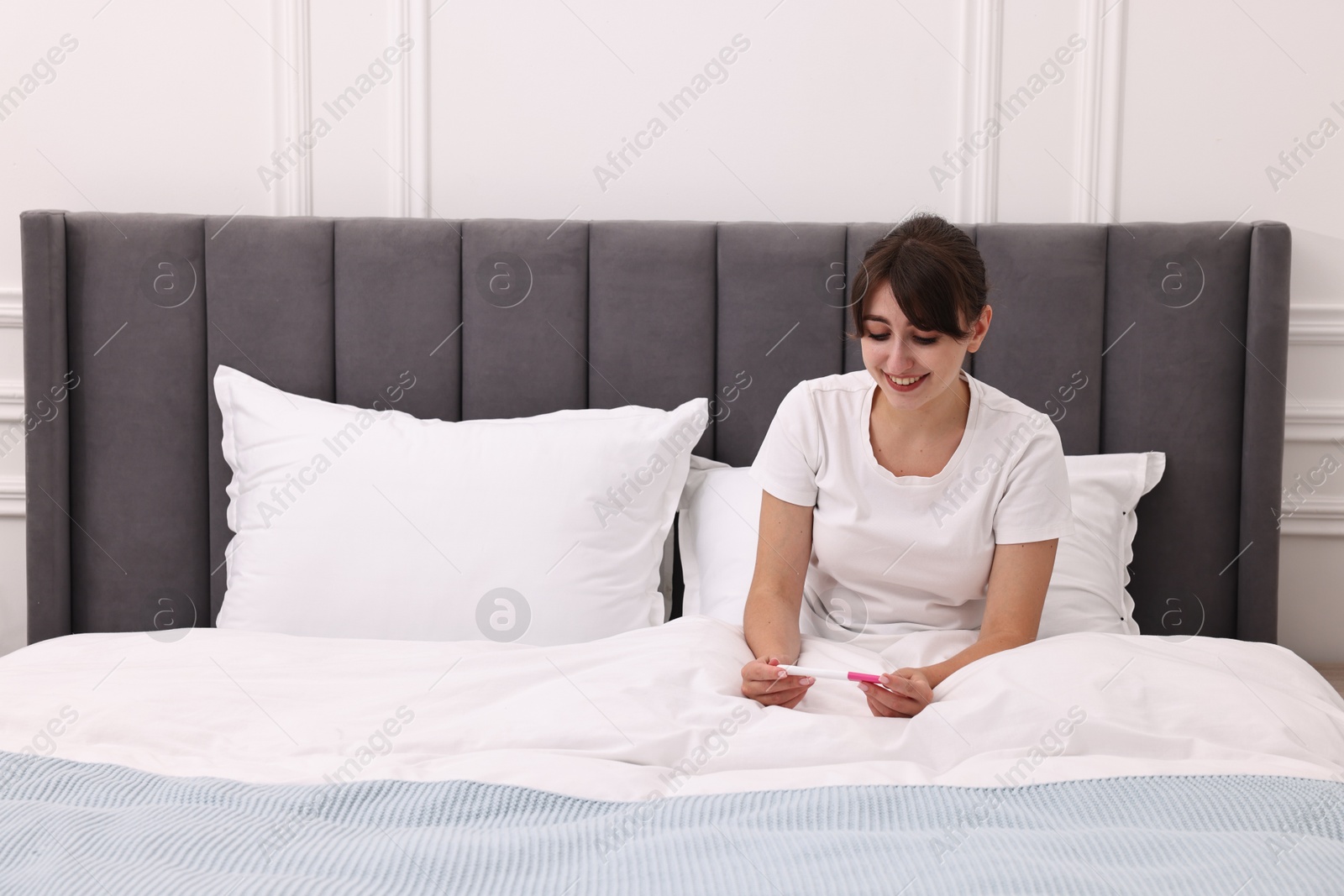 Photo of Happy young woman with pregnancy test on bed at home