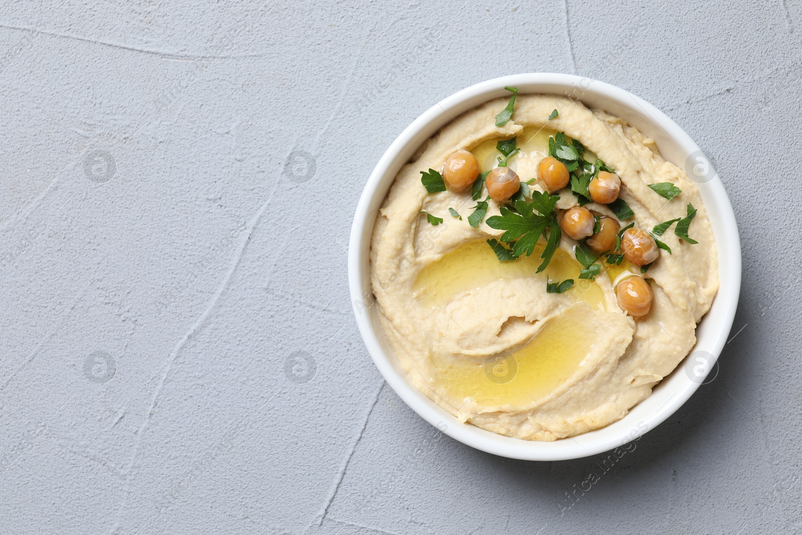 Photo of Delicious hummus with olive oil, chickpeas and parsley in bowl on grey textured table, top view. Space for text