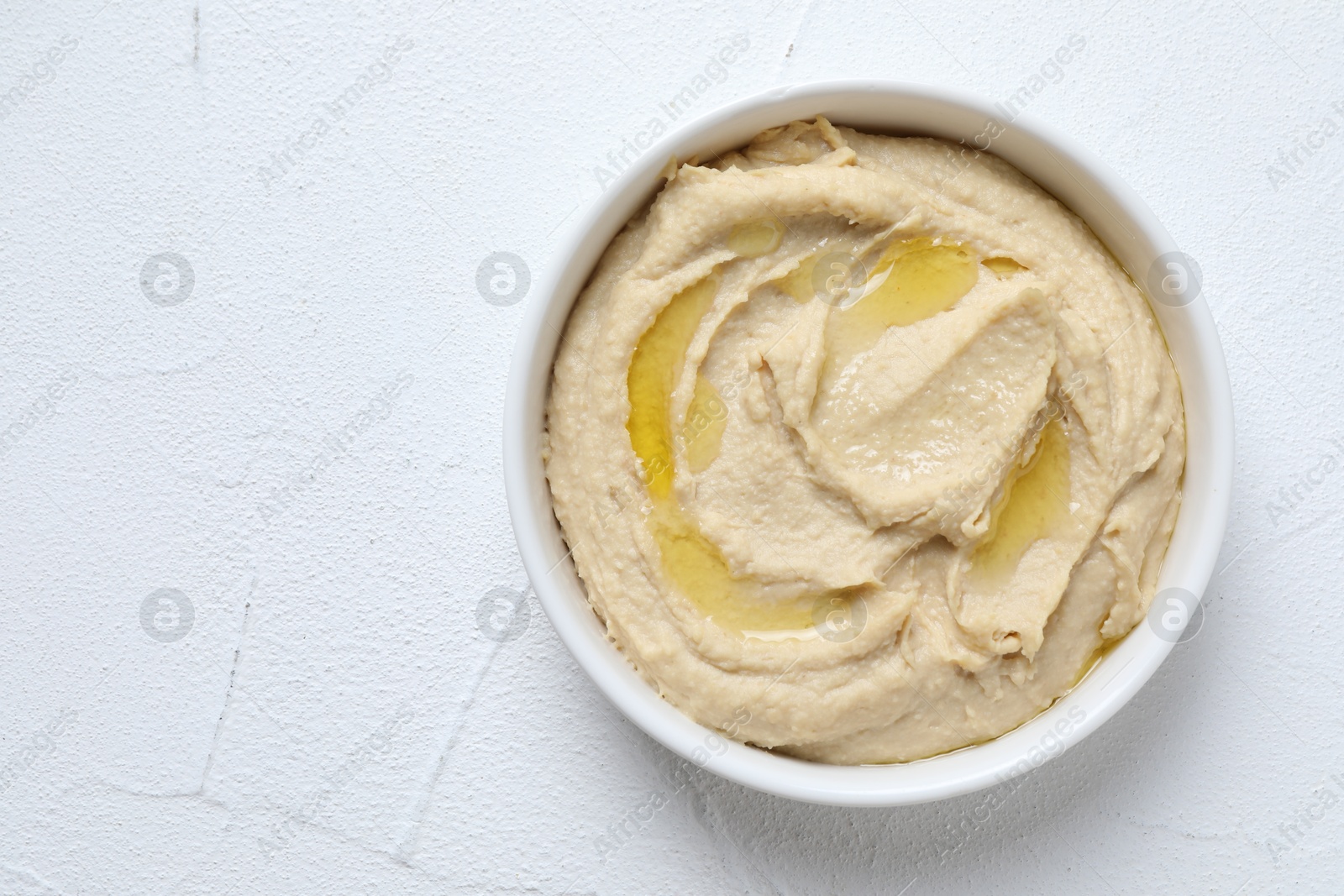 Photo of Delicious hummus with olive oil in bowl on white textured table, top view. Space for text