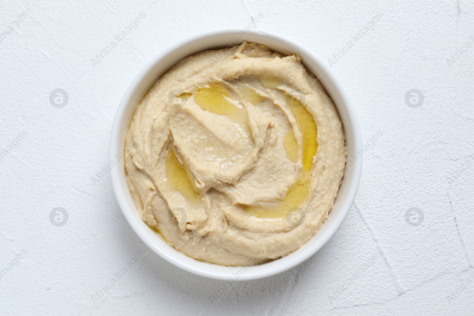 Photo of Delicious hummus with olive oil in bowl on white textured table, top view