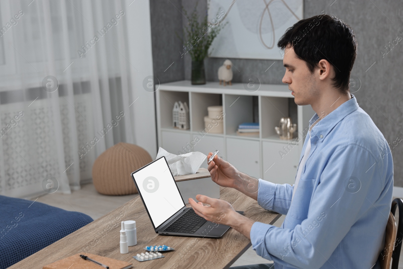 Photo of Sick man with thermometer having online consultation with doctor via laptop at wooden table indoors