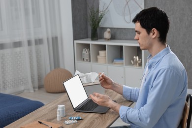 Photo of Sick man with thermometer having online consultation with doctor via laptop at wooden table indoors