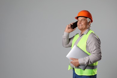 Engineer in hard hat with laptop talking on smartphone against grey background, space for text