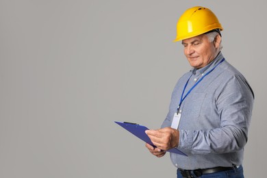 Engineer in hard hat with clipboard on grey background, space for text