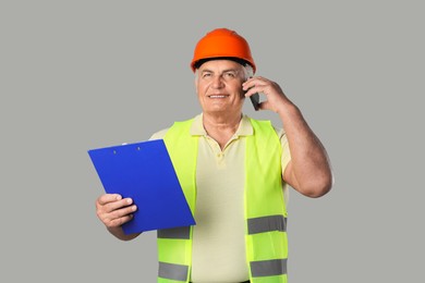 Engineer in hard hat with clipboard talking on smartphone against grey background