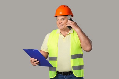 Engineer in hard hat with clipboard talking on smartphone against grey background