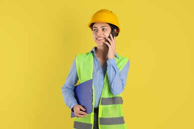 Engineer in hard hat with clipboard talking on smartphone against yellow background