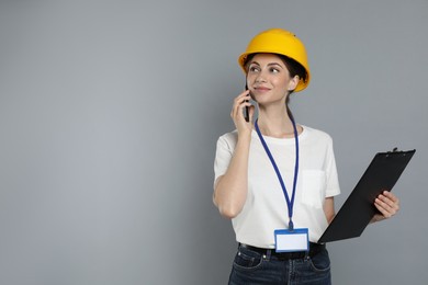 Engineer in hard hat with clipboard talking on smartphone against grey background, space for text