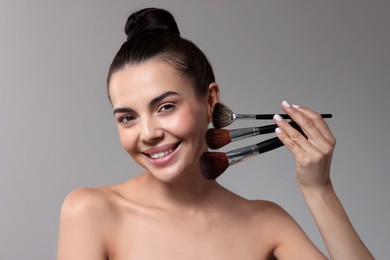 Smiling woman with beautiful makeup holding brushes on light grey background