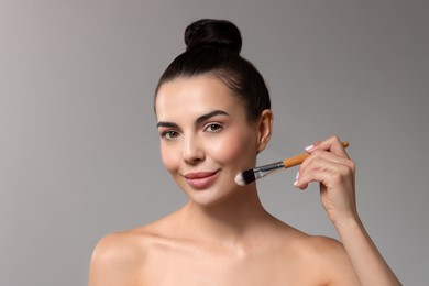 Beautiful woman applying makeup with brush on grey background