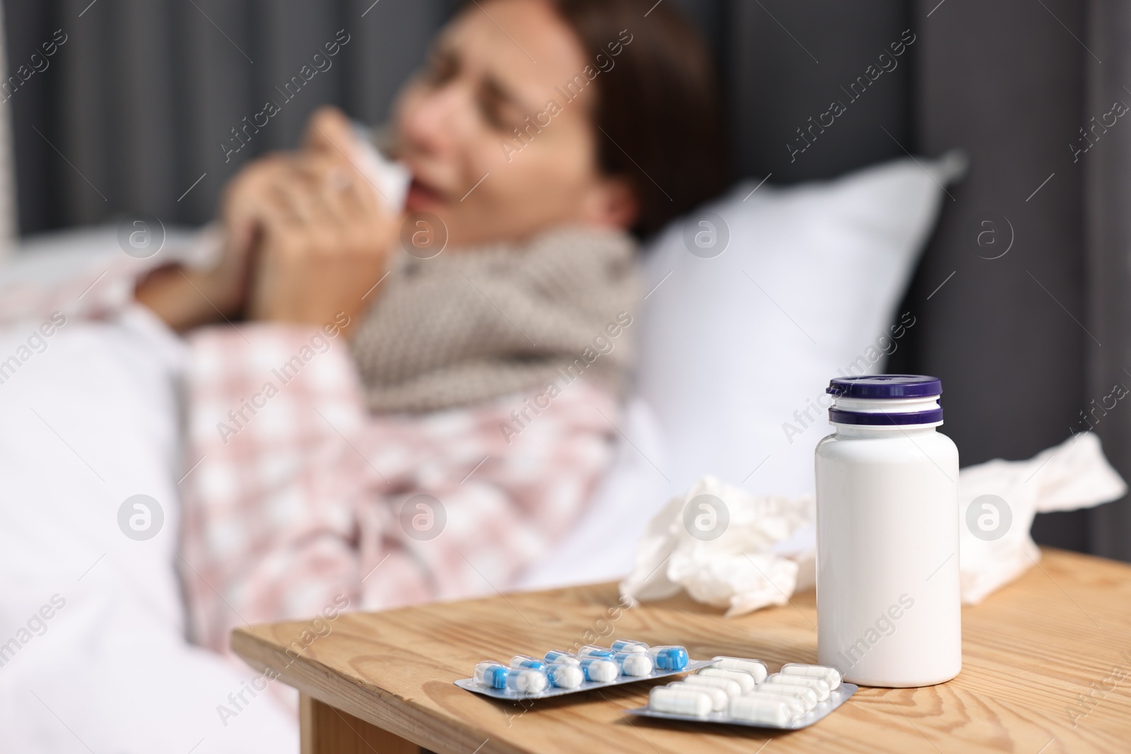 Photo of Sick woman with tissue in bed, focus on different pills on wooden bedside. Cold symptoms