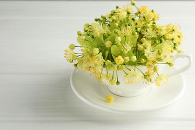 Fresh linden leaves and flowers in cup on white wooden table, space for text