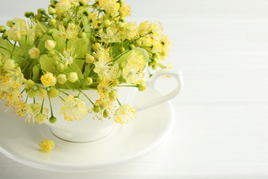 Fresh linden leaves and flowers in cup on white wooden table, space for text