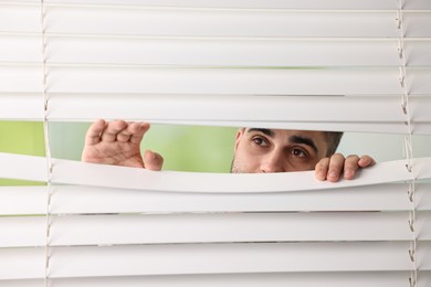 Young man looking through window blinds on blurred background