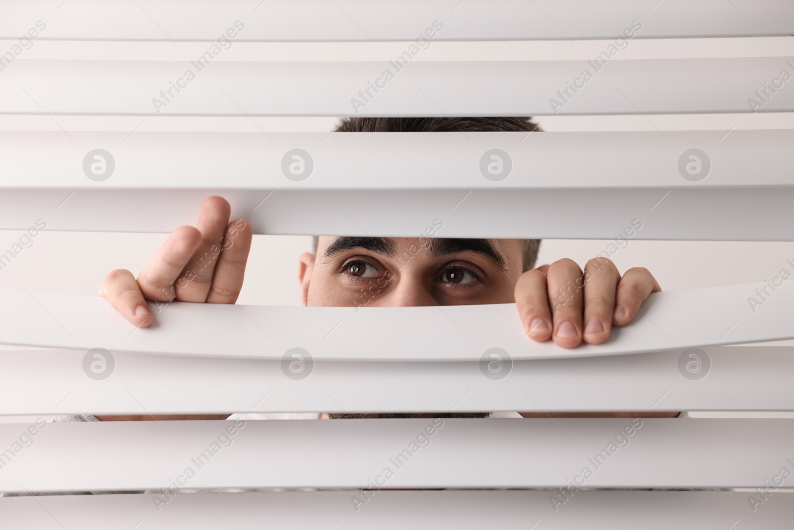 Photo of Young man looking through window blinds on white background