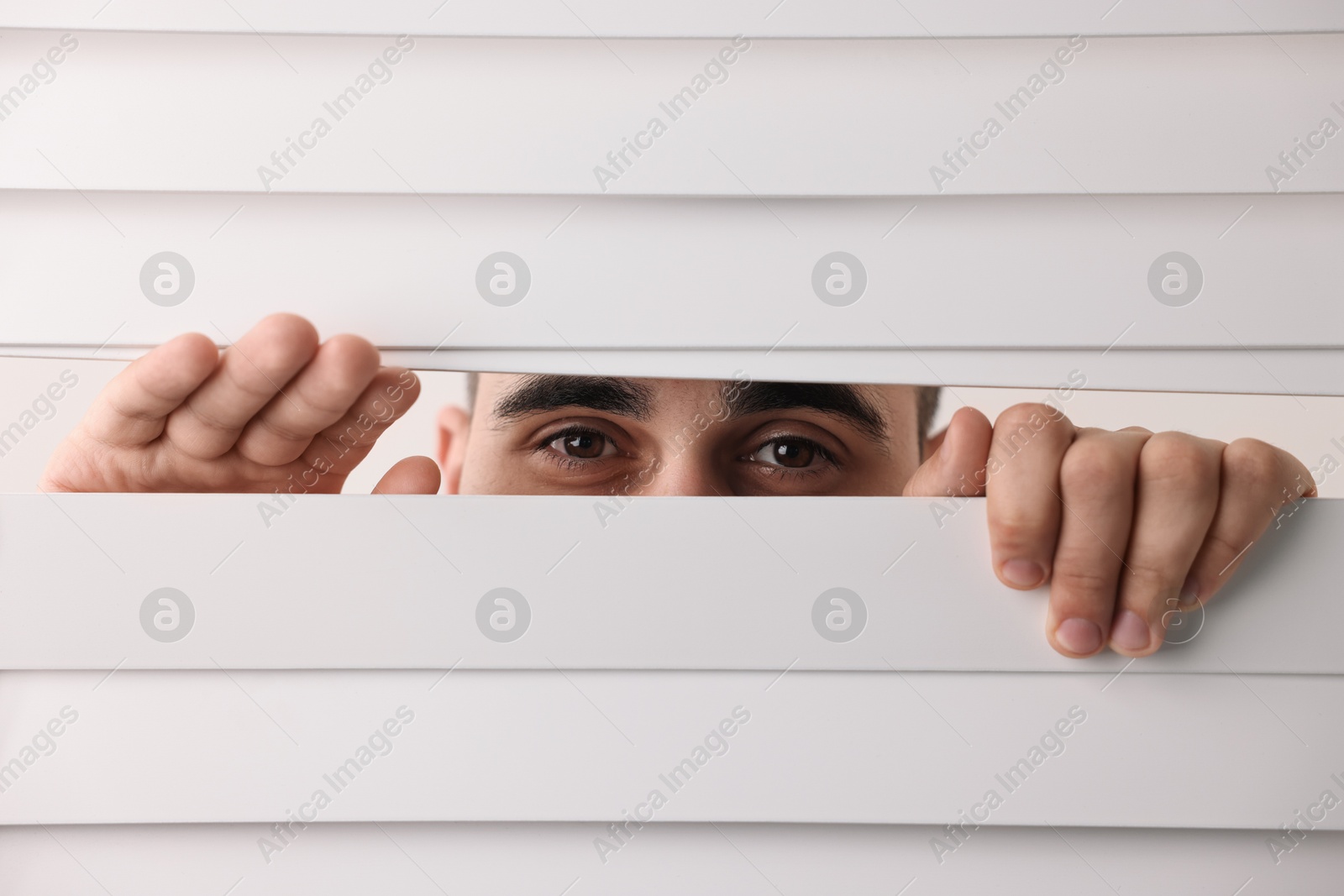 Photo of Young man looking through window blinds on white background