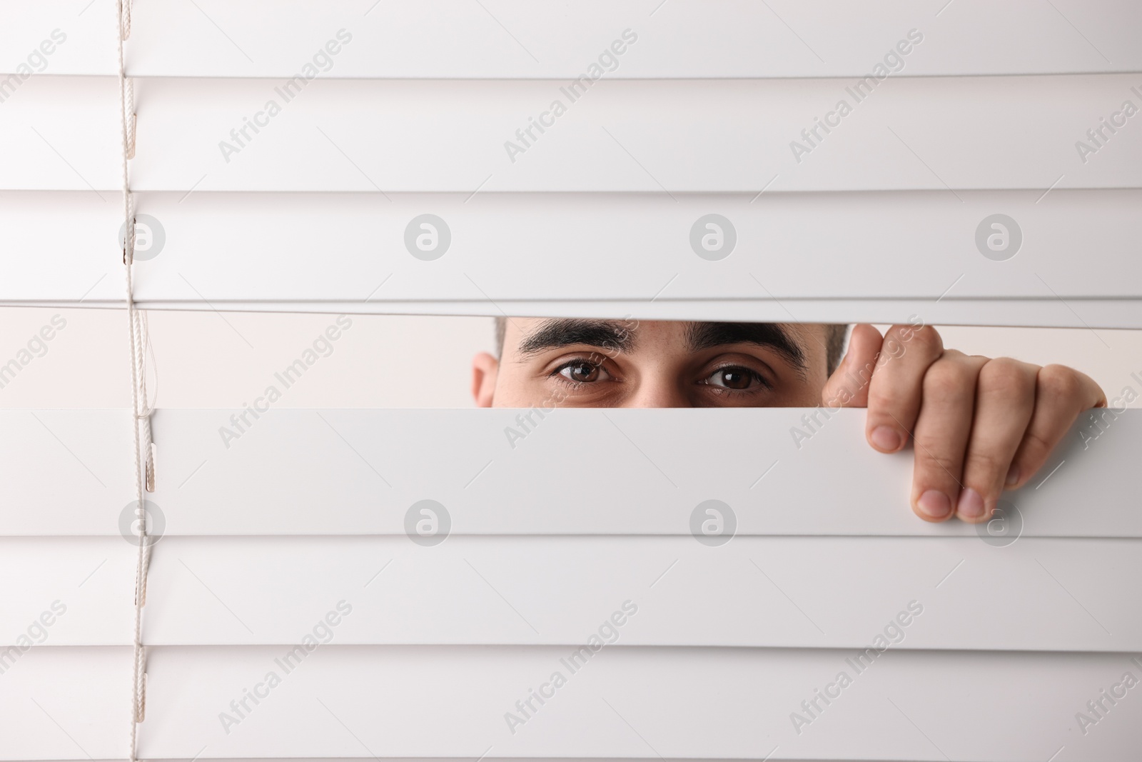 Photo of Young man looking through window blinds on white background