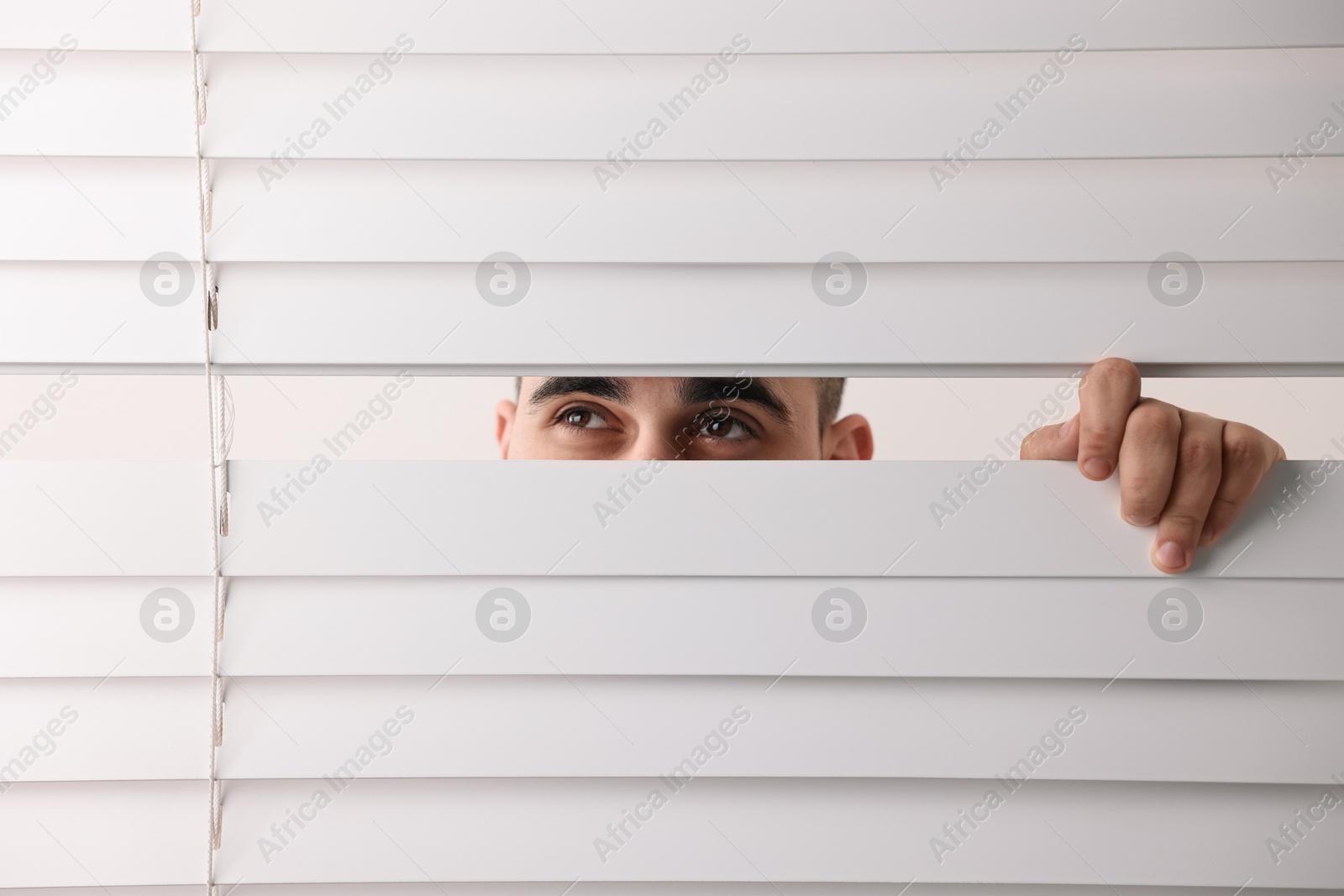 Photo of Young man looking through window blinds on white background