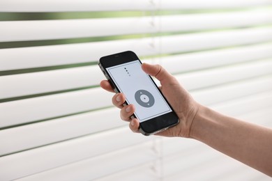 Photo of Woman using smartphone to adjust window blinds indoors, closeup