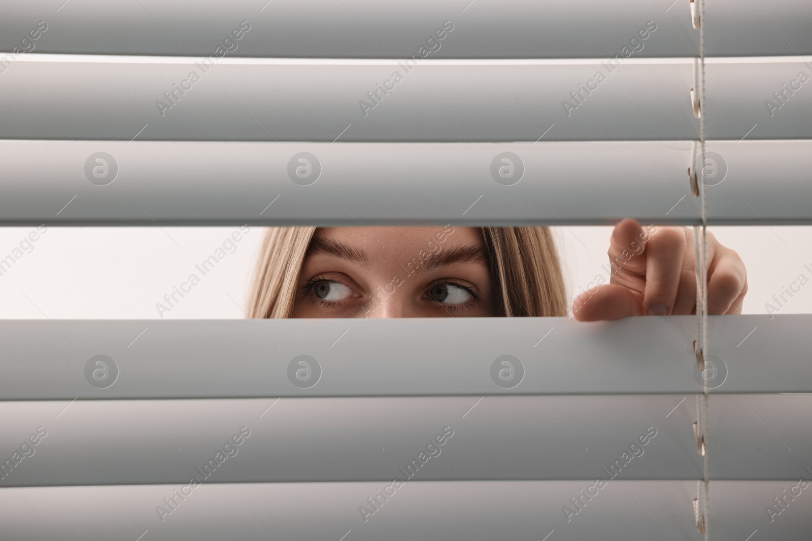 Photo of Young woman looking through window blinds on white background