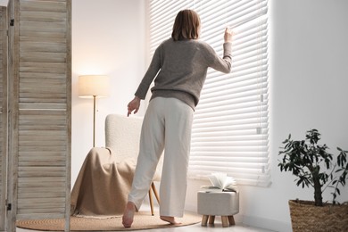 Photo of Young woman near window blinds at home, back view