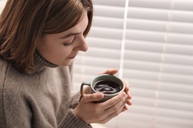 Woman with cup of drink near window blinds indoors, space for text