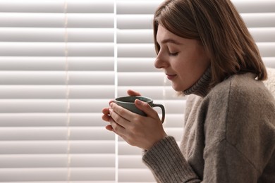 Photo of Woman with cup of drink near window blinds indoors, space for text