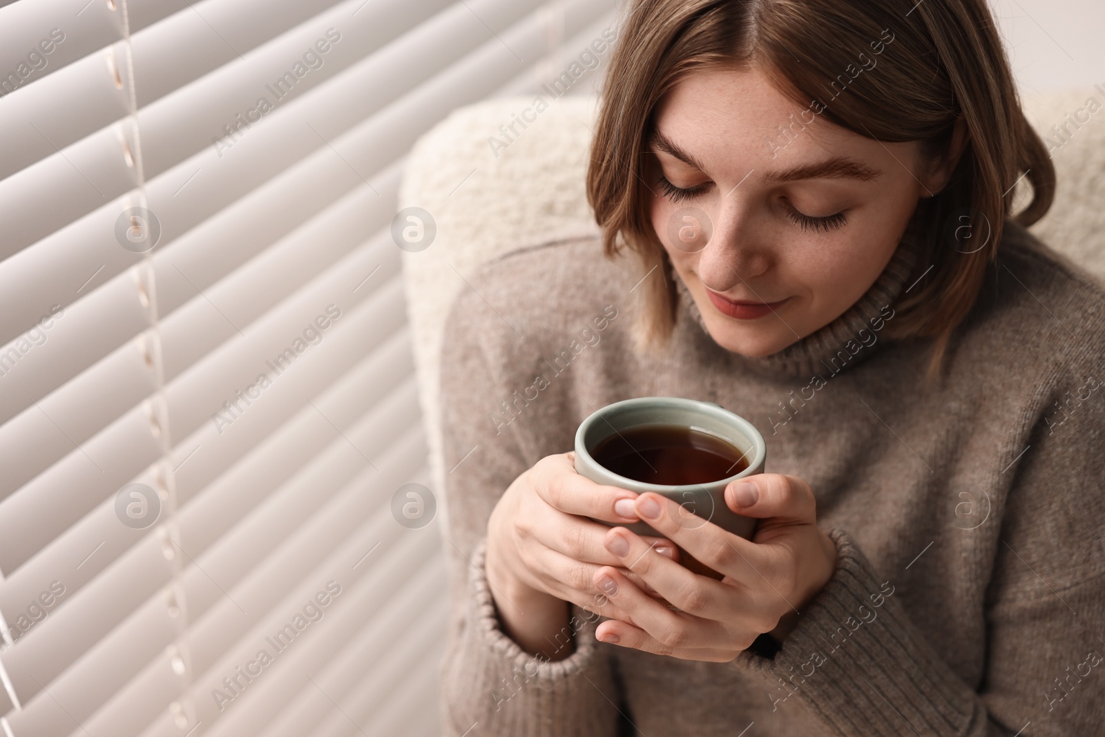 Photo of Woman with cup of drink sitting on armchair near window blinds indoors