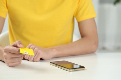 Man holding SIM card near smartphone at white wooden table indoors, closeup