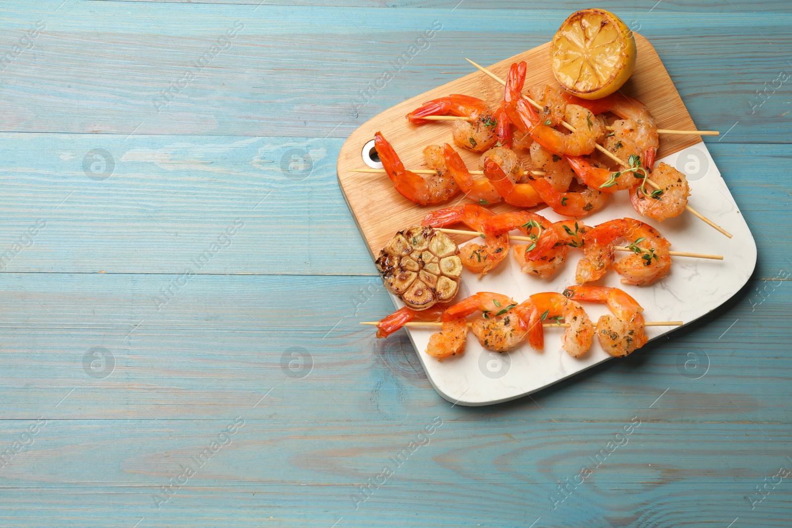 Photo of Delicious grilled shrimp skewers, lemon and garlic on light blue wooden table, top view. Space for text