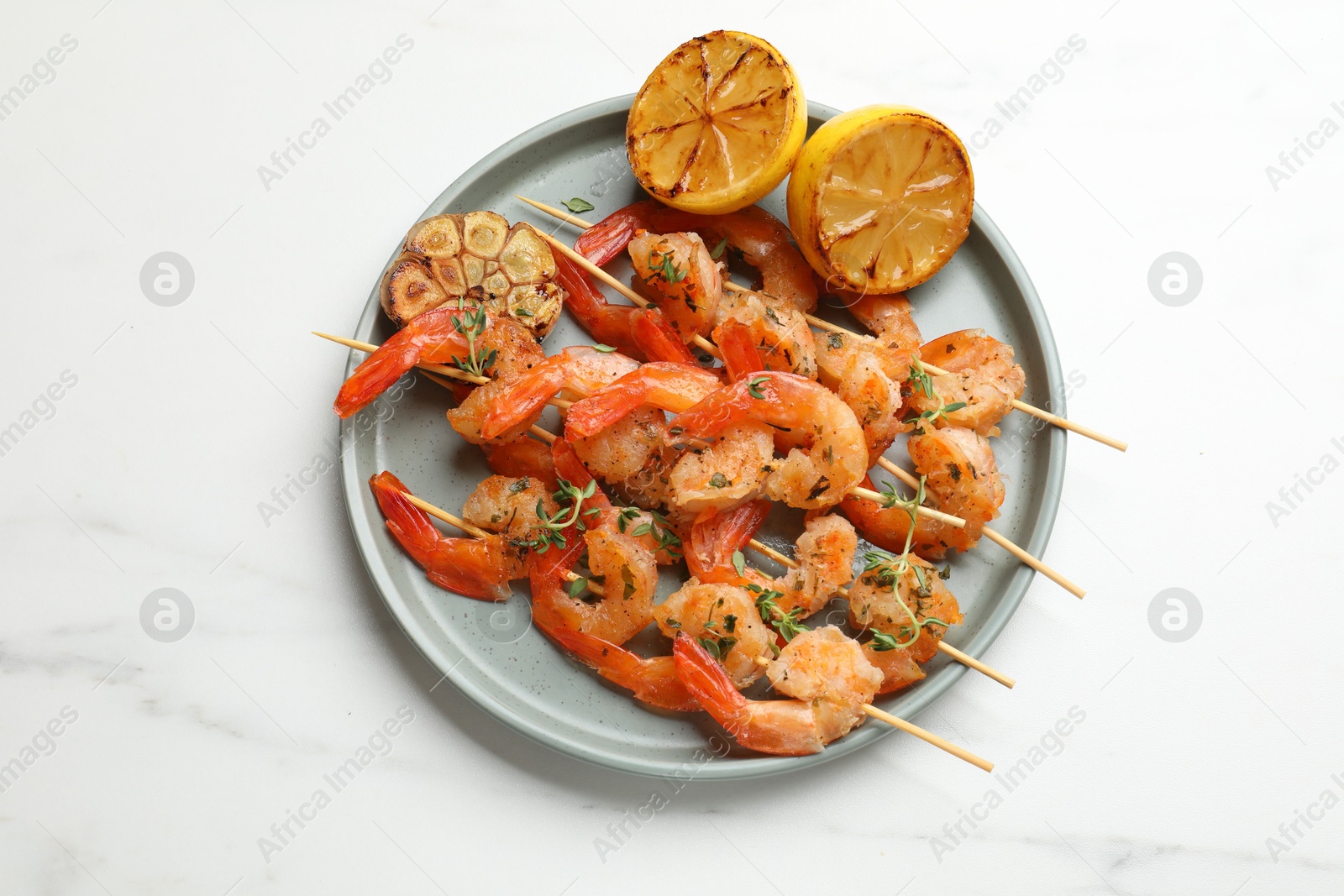 Photo of Skewers with delicious grilled shrimps, lemon and garlic on white marble table, top view