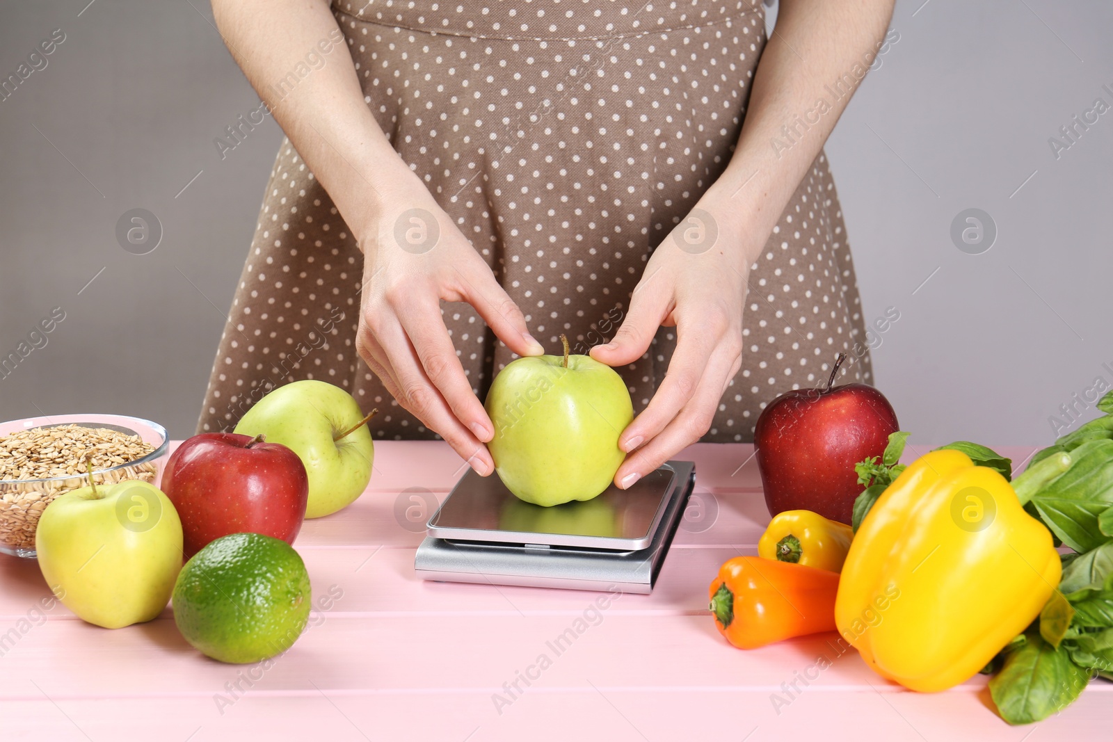 Photo of Woman weighing apple on kitchen scale at pink wooden table, closeup