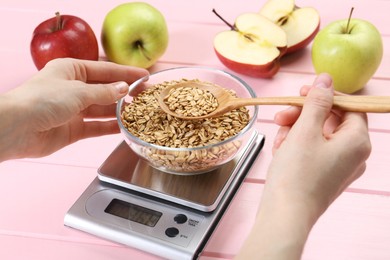 Photo of Woman weighing bowl of oats on kitchen scale at pink wooden table, closeup