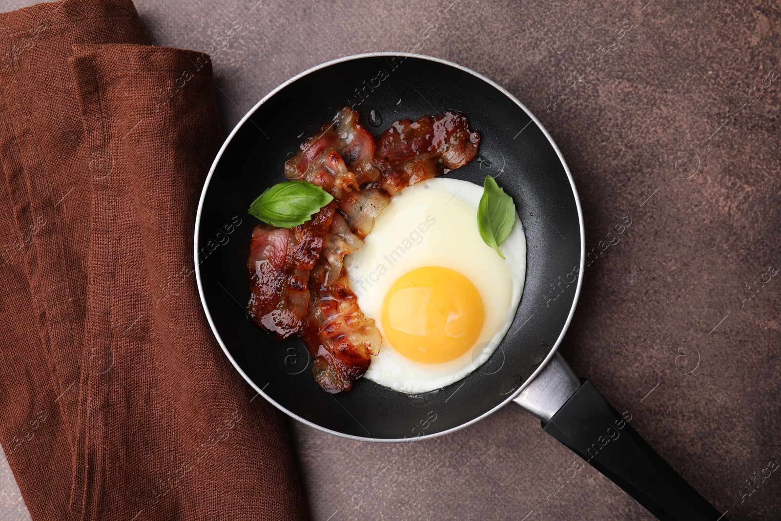 Photo of Fried egg, bacon and basil in frying pan on brown table, top view