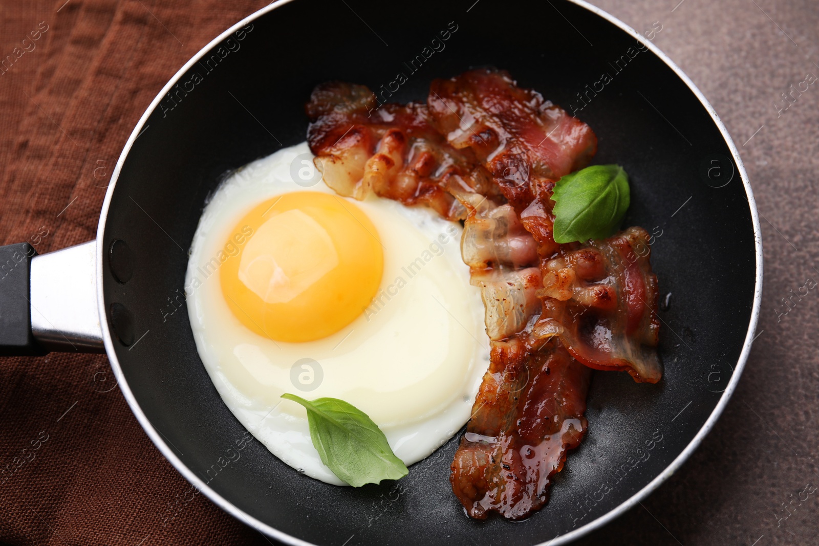 Photo of Fried egg, bacon and basil in frying pan on brown table, above view