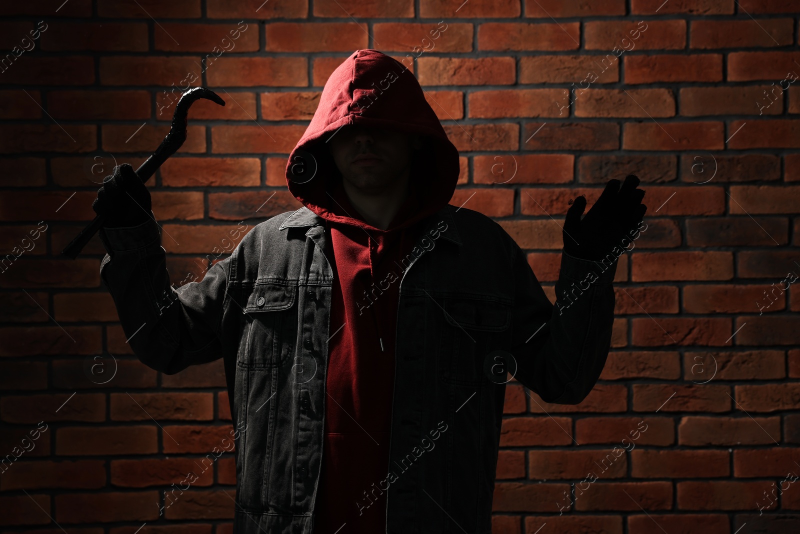 Photo of Thief in hoodie raising hands with crowbar against red brick wall